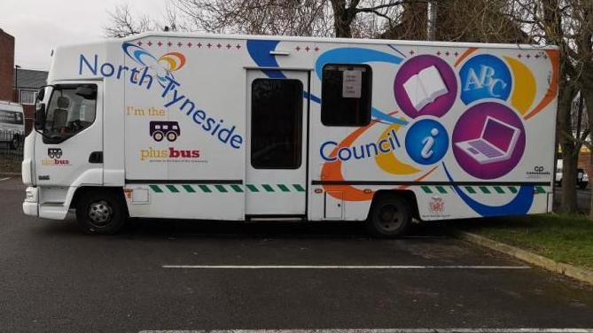 A mobile library, with a central door. It is white, with coloured logos reading: "North Tyneside Council", "I'm the plus bus" and images of an open book and a lap top in purple circles.
