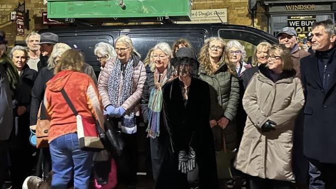 The image depicts a group of people gathered outdoors at night in a town setting. They are standing on a street in front of a row of traditional stone buildings with pitched roofs and multiple windows, some of which are illuminated. Behind the group, there is a dark vehicle with a green make shift coffin on the roof rack, and several shop signs are visible on the buildings. The individuals are dressed in warm clothing, such as coats and scarves, suggesting cool weather with some in black to symbolise a mourning setting. One person in the foreground is wearing a bright red jacket and blue jeans, holding a bag, and a small dog is standing near their feet.