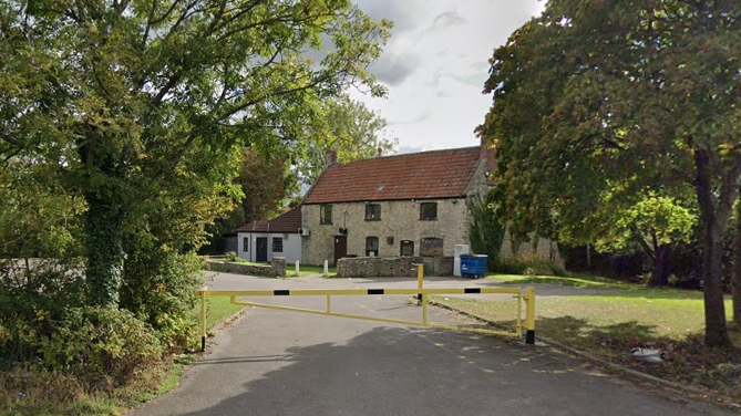 A general view of the Whitchurch Folk House in Bristol, seen from the nearest street. It is framed by trees and has a yellow and blue traffic barrier in front of it. The building itself is made of light brick and has a red tiled roof and black-framed windows