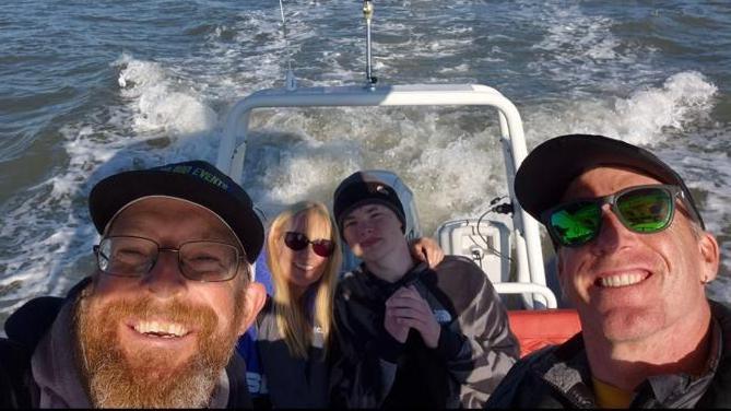 Four people on a speed boat, two men in the foreground with Harrison and a woman to the rear