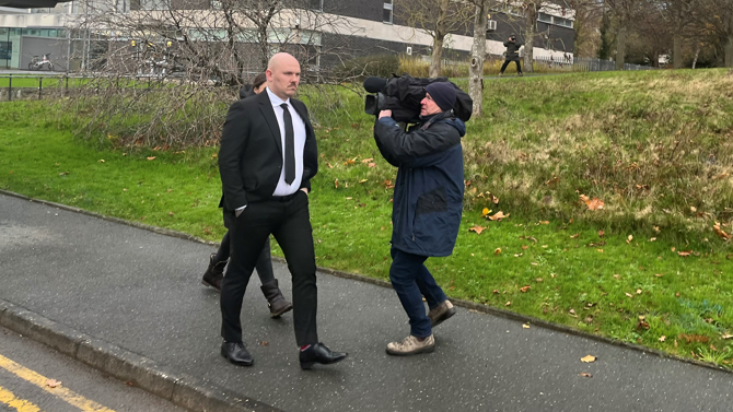 Miles Cross is walking on a pavement, wearing a black suit and tie. In front of him is a camera man pointing a large camera, balanced on his shoulder, at Cross' face.