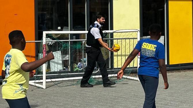 A police officer plays football with some young boys at the Morningside Community Youth Club in Hackney, London. The officer plays in goal and is about to roll the yellow ball out to the four boys, who play on a paved area outside the club, which is painted yellow. 