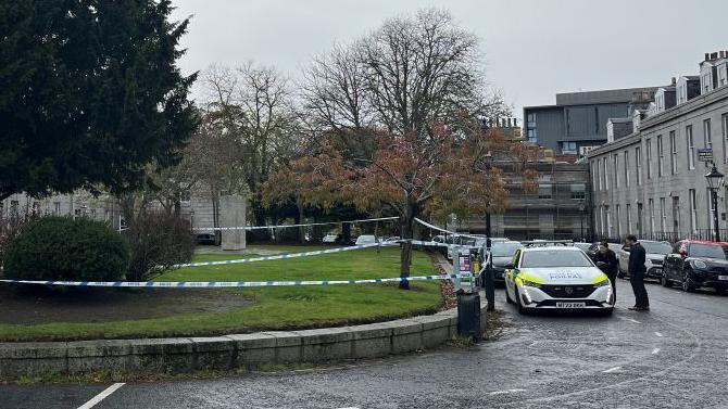 Police tape cordons off a grassy area with trees in a city square, surrounded by parked cars and stone buildings.