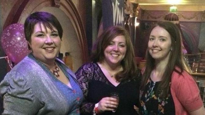 Ciara Hunter has short dark hair and is smiling at the camera with a grey cardigan and blue top with a beaded necklace. Beside her is her sister Joanne who has long brown hair and is wearing a black laced top with a drink in her hand. Next to her is their late sister Clare who has long brown hair a black patterned top and pink cardigan.