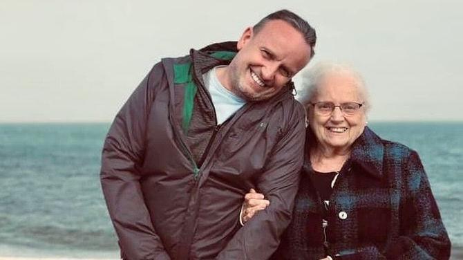 Peter Killian and his mother Lilian smile and hold each other's arms while stood in front of the sea on a beach. Both are wearing coats.