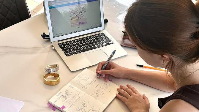 A shot from just above and to the side of Nahla, showing her working on the project. A silver and gold version of the bracelet are on her desk, and she is sketching clasp mechanisms. Her laptop is open in front of her, showing some of the mock-up adverts.