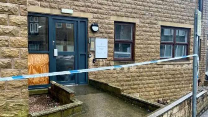 Police tape outside a block of flats on a wet day, with a broken window boarded up. 