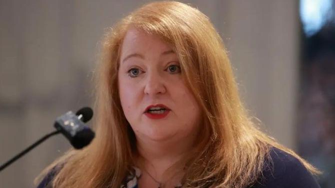 Naomi Long has long red hair and is wearing a purple top. 
She is standing in front of a microphone. 