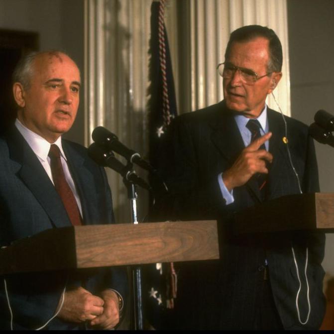 Two men stand infront of podiums with microphones. Both are wearing suits and ties