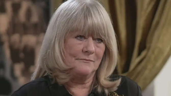 Christine Flack looks to the right of the camera as she is being interviewed. She has a solemn expression and is wearing a dark blouse. She has long blonde hair with a fringe. 