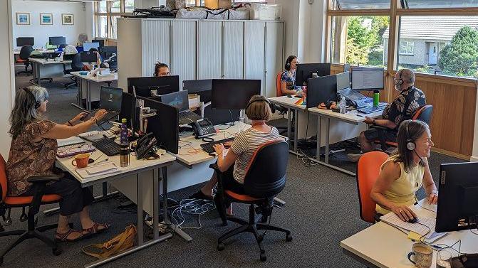 Six women are working in an office. Each of them wears a headset and sits in front of a monitor. There are windows revealing a garden and a building opposite.