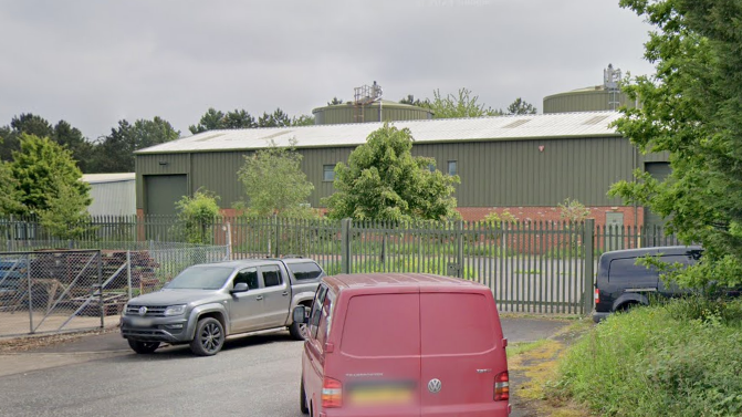 A green corrugated metal building stands behind green metal fences. The car park is empty. In front of the fence and gate are three vehicles