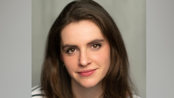 A close-up picture of Clementine Collett. She has long brown hair and brown eyes and is wearing a black and white striped T-shirt. Behind her is a grey background.
