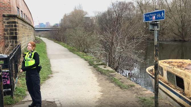 A female police officer wearing a hi-vis tabard stands on the tow-path by the River Aire, next to an old, rusty boat.