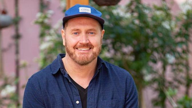 A man with a ginger beard wearing a blue baseball cap, black t shirt and blue shirt and smiling at the camera. He is sat in front of some green plants.