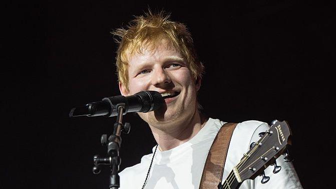 Ed Sheeran performs on stage. He stands in front of a microphone and looks away from the camera while smiling. He has red hair and wears a white T-shirt. A guitar strap rests around his torso as he plays.