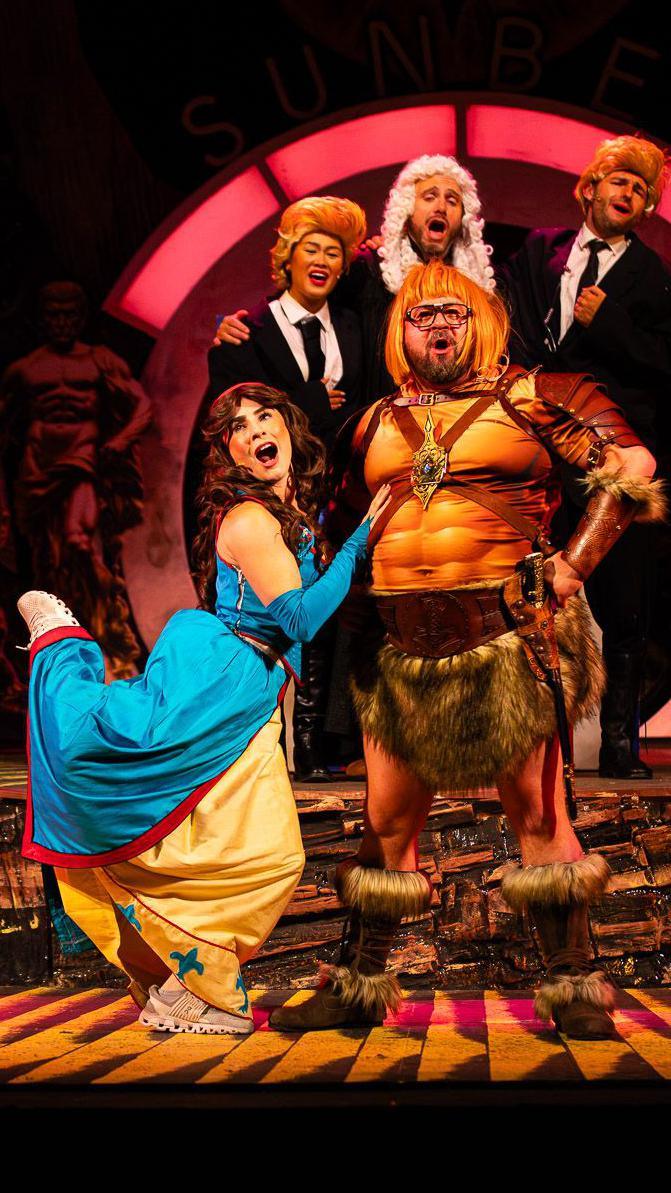 Stage photograph of actors in medieval clothes playing Coleen and Wayne Rooney, with three actors in suits and judges' wigs behind them