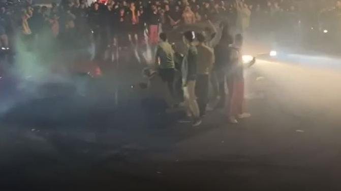 Two cars with headlights on driving around a circle of people at night. You can see the cars' lights beaming into the crowd of people.