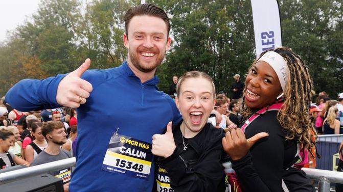 Group picture of Calum Lill in a blue top, and Harriet Bibby and Channique Sterling-Brown who are wearing black tops near the starting point. They are all grinning and giving thumbs-up signs. Other runners are behind them as they wait to start.