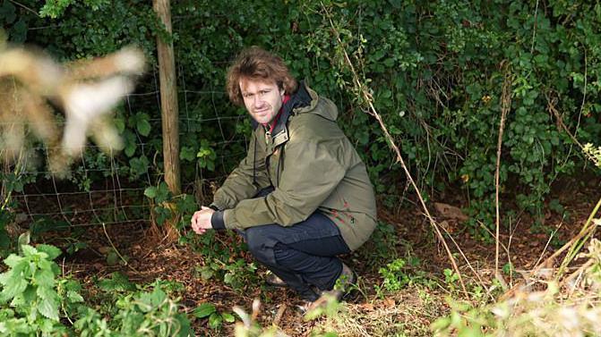 A person in an olive-green outdoor jacket crouches beside a wire fence at the edge of dense woodland. The ground is covered with leaves, grass and low vegetation, while thick green foliage fills the background. Softly blurred plants frame the foreground, drawing attention to the secluded countryside setting.