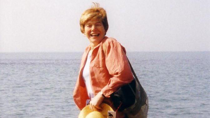 Woman wearing orange shirt, with bag over one shoulder, holding a hat, while smile on the beach, with the sea behind her