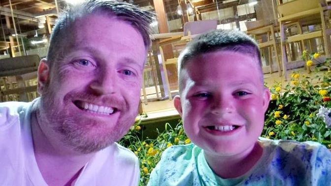 James Askew in a white T-shirt sits next to his son Alex in a green and blue speckled T-shirt