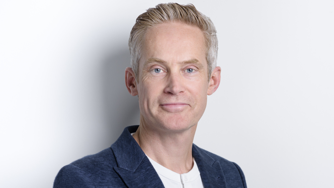 Guy Collis looking directly at the camera. He is stood against a white wall or background. He is wearing a blue suit jacket over a white t-shirt. 