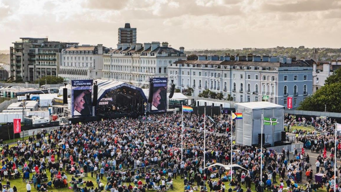 Aerial view of a large crowd at summer sessions in Plymouth. Large crowds have gathered on the Hoe to watch artists on a large stage. Residential properties are located behind the stage.
