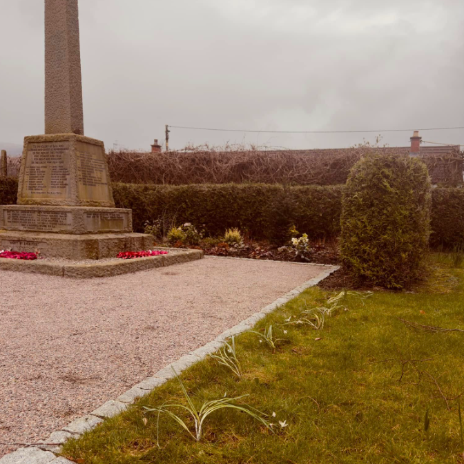 A war memorial with a gravel path. Beside it is a grassy area where snowdrops have ben planted