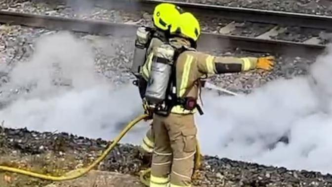 Two firefighters wearing beige outfits and silver back cannisters and illuminous helmets hold a water pipe over the smoke on the rail tracks.