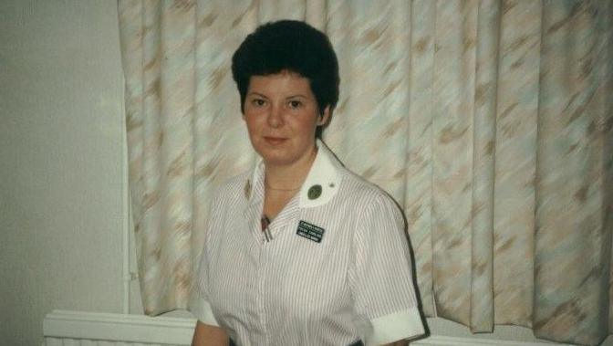 Woman in a nurse's uniform, with short hair and a striped dress and blue belt, standing in front of curtains and a radiator