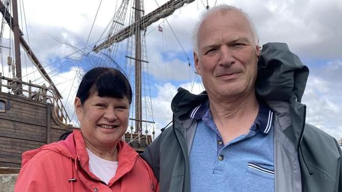 Dawn and James Paine standing outside in front of a wooden ship. Dawn Paine is wearing a white T-shirt and red coat and is looking directly at the camera and smiling. James Paine is wearing a blue T-shirt and grey coat.
