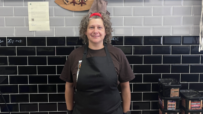 Lilith is standing against black tiles in her shop on the high street. She has brown curly hair pulled back with a red band and a black apron on over a brown T'shirt.