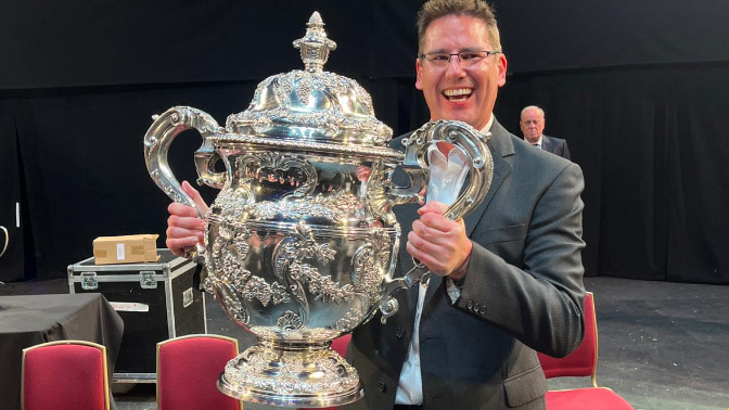 Paul Holland smiling to camera holding the enormous silver trophy in his arms