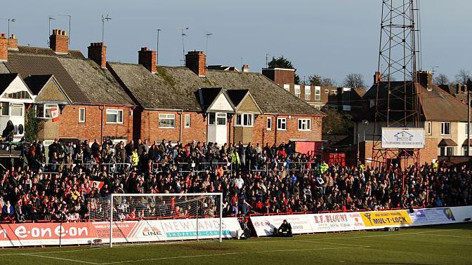 Fans packed into one of the stands behind the goal at Rockingham Road before a game. Behind the stand houses overlooking the ground are visible, as well as the base of a large floodlight.