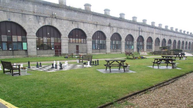 HMP The Verne building in the back of the picture, with a grassy area in front of it. The building is a one-storey building with turrets at regular intervals built onto the roof. There are picnic benches and a large-scale chess board and pieces on a board paved on the ground. It appears to be a wet or misty day when the picture was taken