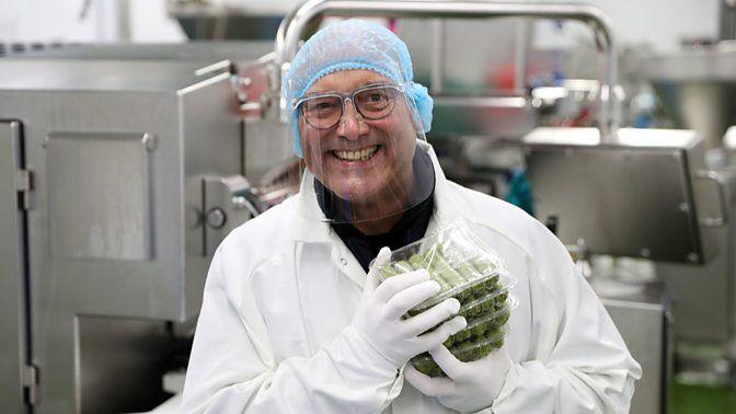 Wallace, inside a factory that produces vegan sausages, wearing a blue hairnet, glasses, visor, white lab coat and white gloves. He is holding four packets of sausages