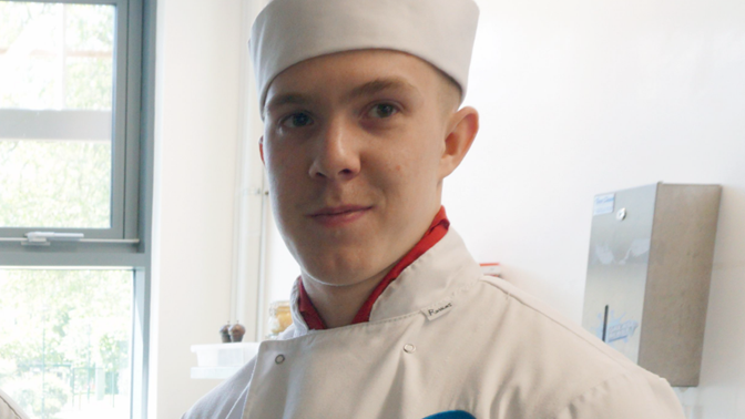 A head and shoulders shot of Joe dressed in white catering clothing and hat. He has a red shirt on underneath