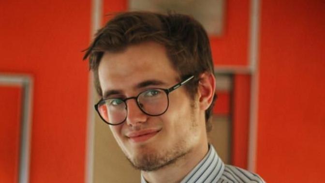 Brad Wright smiles at the camera while standing inside the Clacton Express' passenger carriage. He has short brown hair and is wearing black round glasses and a white shirt with dark stripes. 