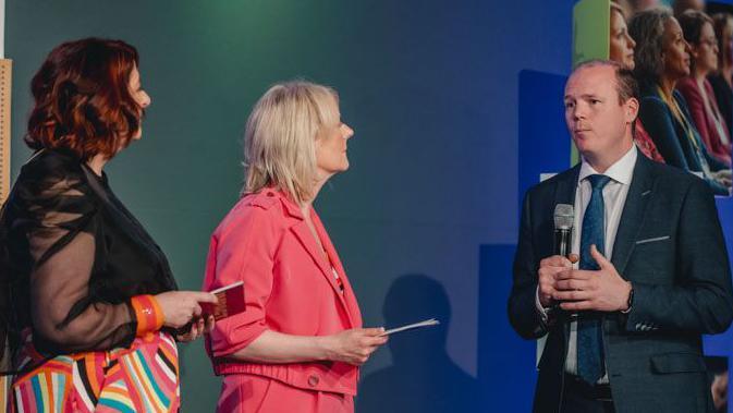 Three people on a stage. A woman on the left has shoulder length red hair. She is looking away from the camera. She is wearing a black mesh shirt and colourful stripy trousers. In the middle a woman with blonde hair is looking at the minister. She is wearing a pink matching suit. On the right, the minister is wearing a grey suit and a blue tie. He has short brown hair and is holding a microphone.