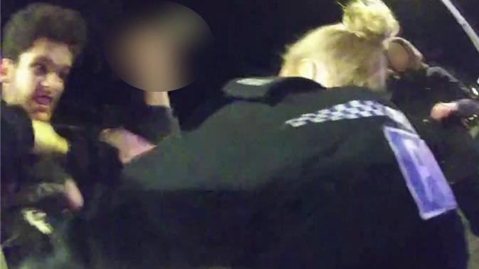 A still image from body-worn police camera footage, showing a female police officer with blonde hair and her black police-issue jacket, who appears to be wrestling with Declan Diedrick, who has thick hair and is gritting his teeth menacingly.