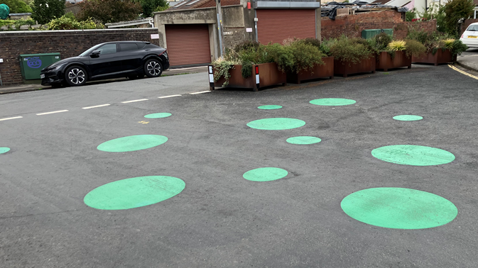A cluster of green spots of various sizes are painted onto a road surface. A large planter full of foliage can be seen behind them. There are parked vehicles to either side.