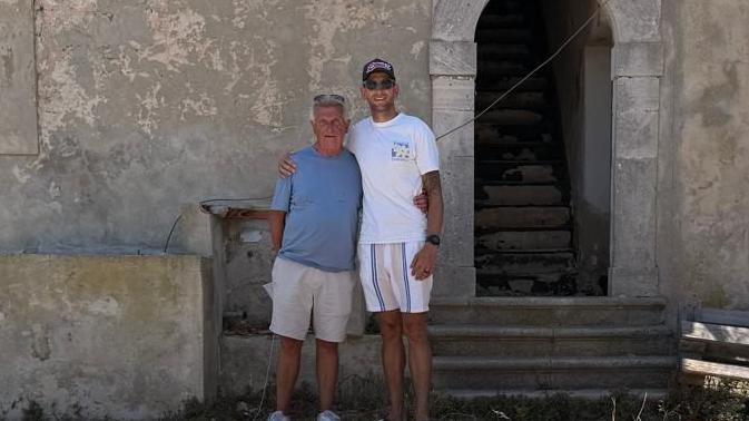 Neal Latham, left, with his youngest son. Mr Latham has a blue t-shirt on with shorts, his son who is taller than him is wearing a white t-shirt with a cap and sunglasses on. They are standing near an old stone wall of a battery in Augusta, Sicily. There is an open stone archway to their left and an two open windows either side.