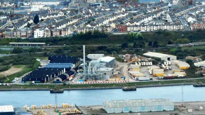 An aerial shot of Barry Docks: in the foreground is a body of water, behind that waste wood incinerator in and amongst industrial equipment. In the background, rows of houses in Barry