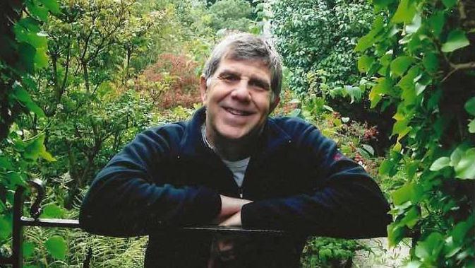 Bill Batcock smiles wearing a fleece as he leans with his arms folded over a black garden fence. A verdant garden can be seen behind him, with trees and shrubs in the distance. 