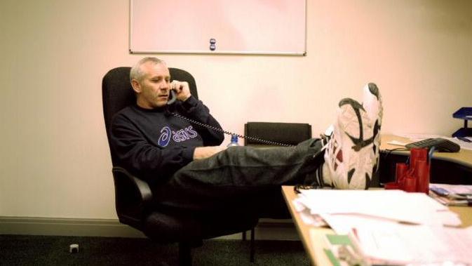 Peter Reid sitting on an office chair with his feet up on his desk, next to several documents. He is wearing a dark sweatshirt with the brand name asics on the front. 