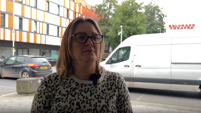 Sarah Windrum standing in the street wearing a leopard print t-shirt. She is wearing a microphone and there is a van behind her. 