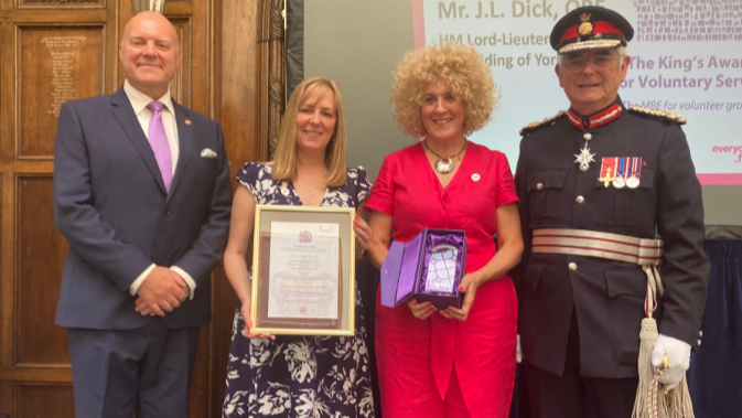Chris Blacksell, Deputy-lieutenant for East Riding of Yorkshire, Vickie Hillier and Sam Barlow, Fitmums & Friends and Mr James Dick OBE, Lord Lieutenant of East Riding of Yorkshire stand in a line. Vickie Hillier is holding a framed certificate and Sam Barlow is holding a blue box.