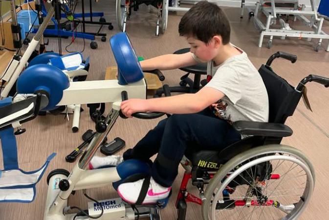 Harry is seen in a wheelchair using a physical therapy exercise machine as part of a rehabilitation room with other equipment in the background. 