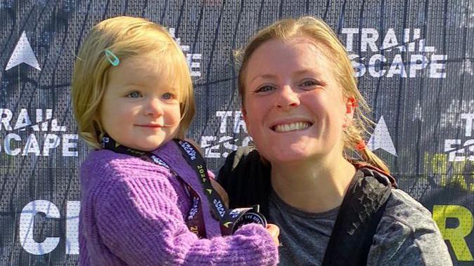 Laura's head and shoulders on he right, smiling at the camera while wearing a grey top and a backpack, while holding her daughter on her hip, who is wearing a purple jumper and her mother's medal.